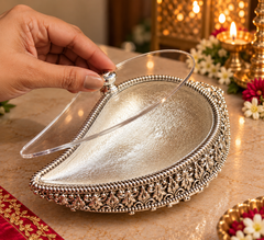 A silver teardrop-shaped decorative tray with ornate filigree detailing around the edge and a clear acrylic lid, shown on a marble surface with a hand lifting the lid. Warm candlelight and floral decorations are visible in the background.
