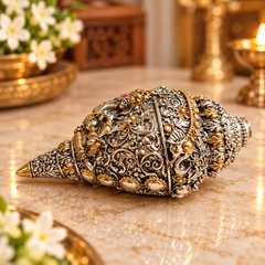 An ornate silver and gold conch shell decorative piece with intricate filigree detailing, displayed on a polished marble surface with warm, blurred background elements.