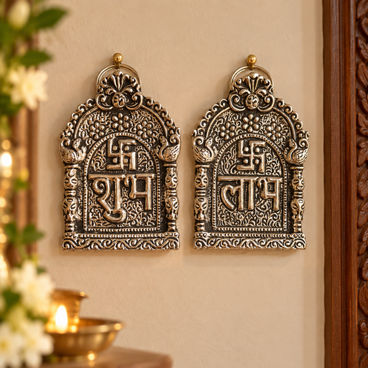 Two ornate metallic wall hangings with carved decorative patterns and Devanagari script, mounted on a beige wall with warm ambient lighting and floral decor partially visible.