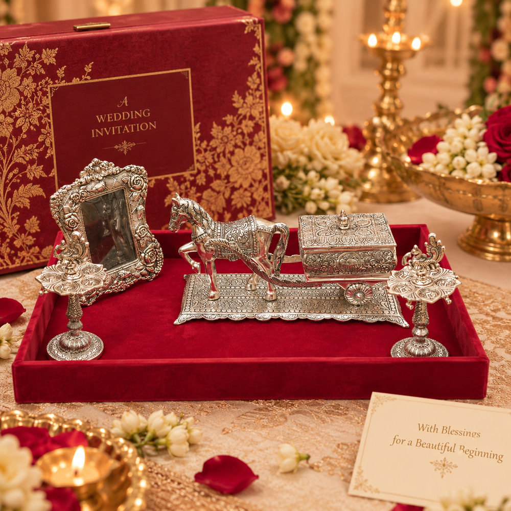 An ornate silver horse-drawn carriage wedding invitation set displayed in a red velvet tray, with a matching red box labeled 'A WEDDING INVITATION' in the background and a decorated wedding table setting around it.