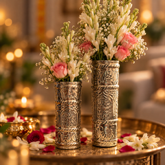Two ornate silver-toned carved vases filled with white baby's breath and pink roses, displayed on a gold-toned table with warm blurred background lights.