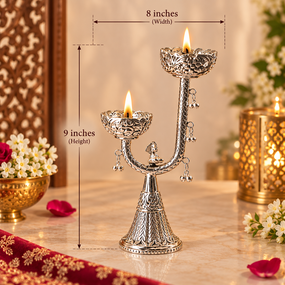 An ornate silver diya holder with two lit oil lamps, shown on a marble surface with measurement overlays indicating 9 inches in height and 8 inches in width. The background features warm bokeh lights, flowers, and decorative elements.