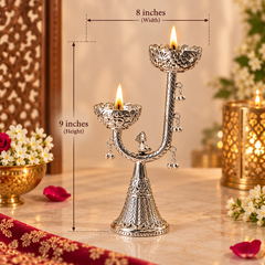 An ornate silver diya holder with two lit oil lamps, shown on a marble surface with measurement overlays indicating 9 inches in height and 8 inches in width. The background features warm bokeh lights, flowers, and decorative elements.
