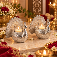 Two silver peacock-shaped candle holders with lit tealight candles on a marble surface, surrounded by flowers and warm decorative lighting in an indoor setting.