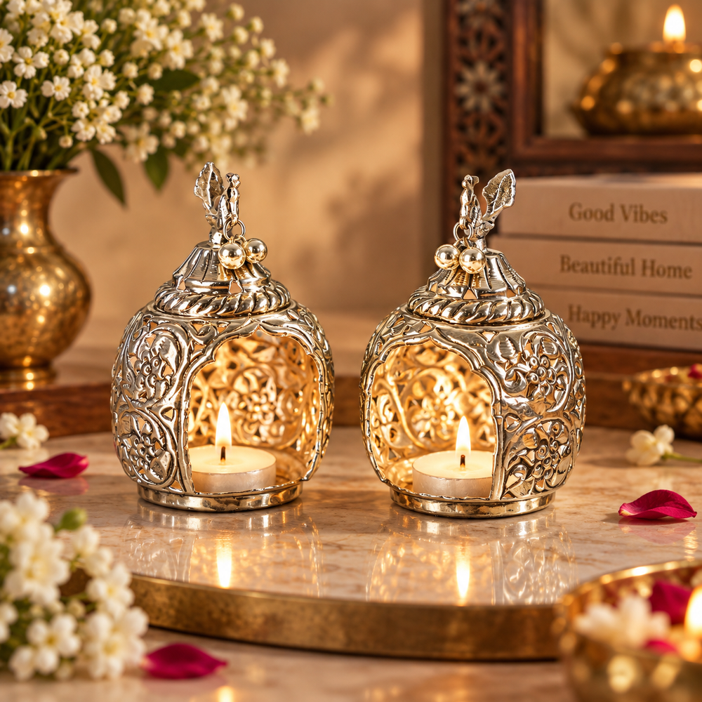 Two ornate silver-toned metal candle lanterns with floral cutout patterns and small decorative finials, each holding a lit tealight candle, displayed on a marble surface with warm decorative props in the background.