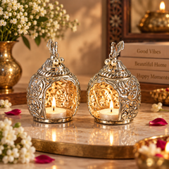 Two ornate silver-toned metal candle lanterns with floral cutout patterns and small decorative finials, each holding a lit tealight candle, displayed on a marble surface with warm decorative props in the background.