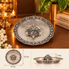 An ornate silver-toned metal tray with a raised turtle motif in the center, shown on a wooden table with decorative candles, flowers, and books. A smaller inset at the bottom includes dimension markings: 15 cm across the tray and 3 cm for the turtle topper.