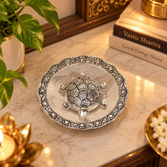 A silver-toned metal turtle figurine sits in the center of an ornate scalloped silver dish on a marble countertop, styled with a potted plant, books, and warm ambient lighting.