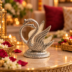 A decorative silver-toned swan figurine with two curved necks and detailed feather patterns, displayed on a reflective gold-toned table in a warmly lit indoor setting with flowers and candles.