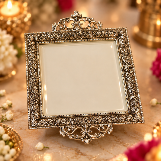 An ornate square silver-toned picture frame with intricate floral scrollwork and a decorative top and bottom crest, displayed on a marble surface with warm candlelight and flowers in the background.