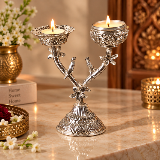 An ornate silver-toned metal candle holder shaped like a flowering branch, holding two lit tealight candles on a marble tabletop in a warmly lit indoor setting.
