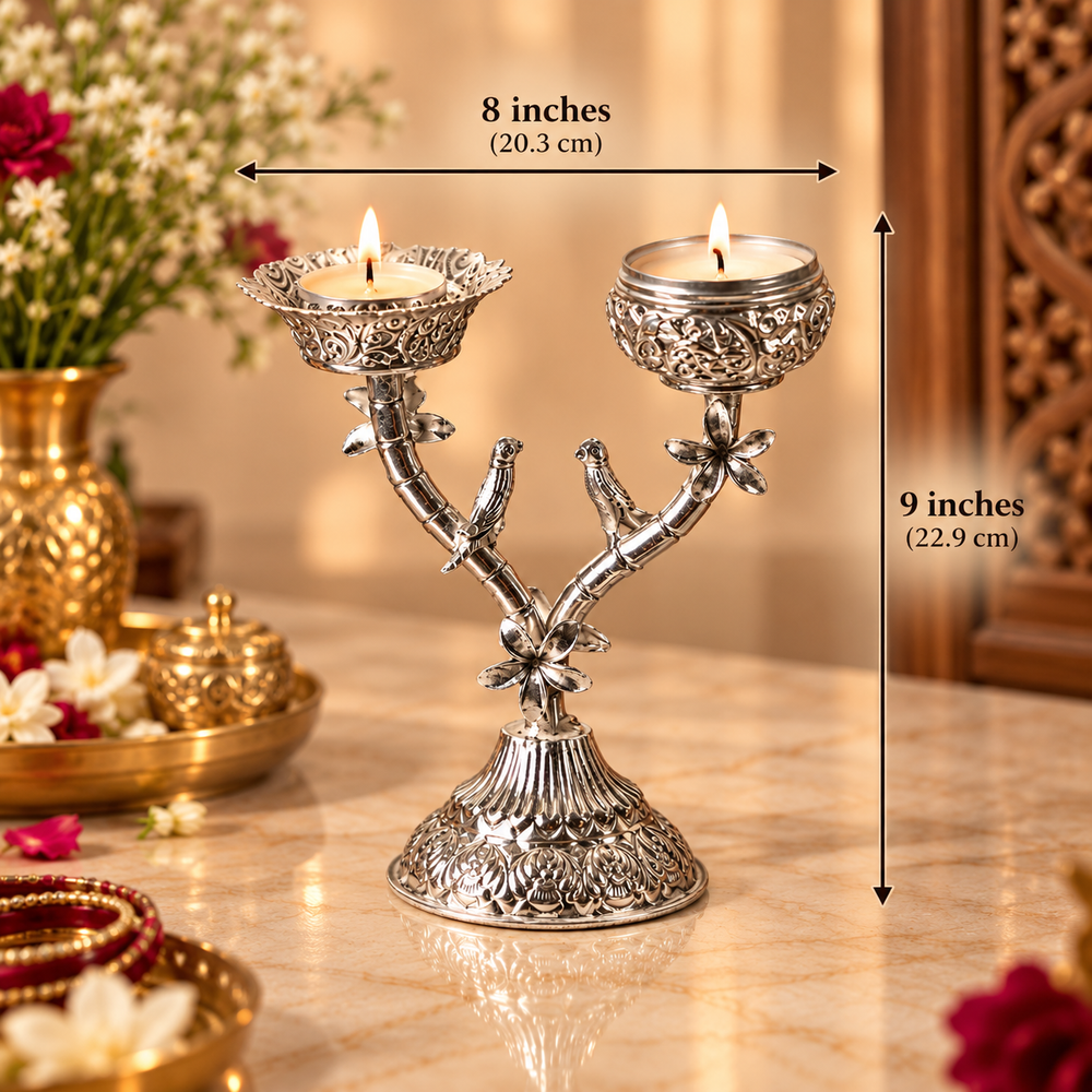 An ornate silver-toned double candle holder with bird motifs on the stem, shown lit on a marble surface with decorative flowers and brass vessels in the background. Black measurement arrows and text indicate 8 inches wide and 9 inches tall.