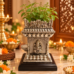 An ornate silver-toned decorative planter shaped like a miniature temple, filled with green leafy stems, displayed on a dark wooden base on a marble surface with warm ambient lights and floral decor in the background.