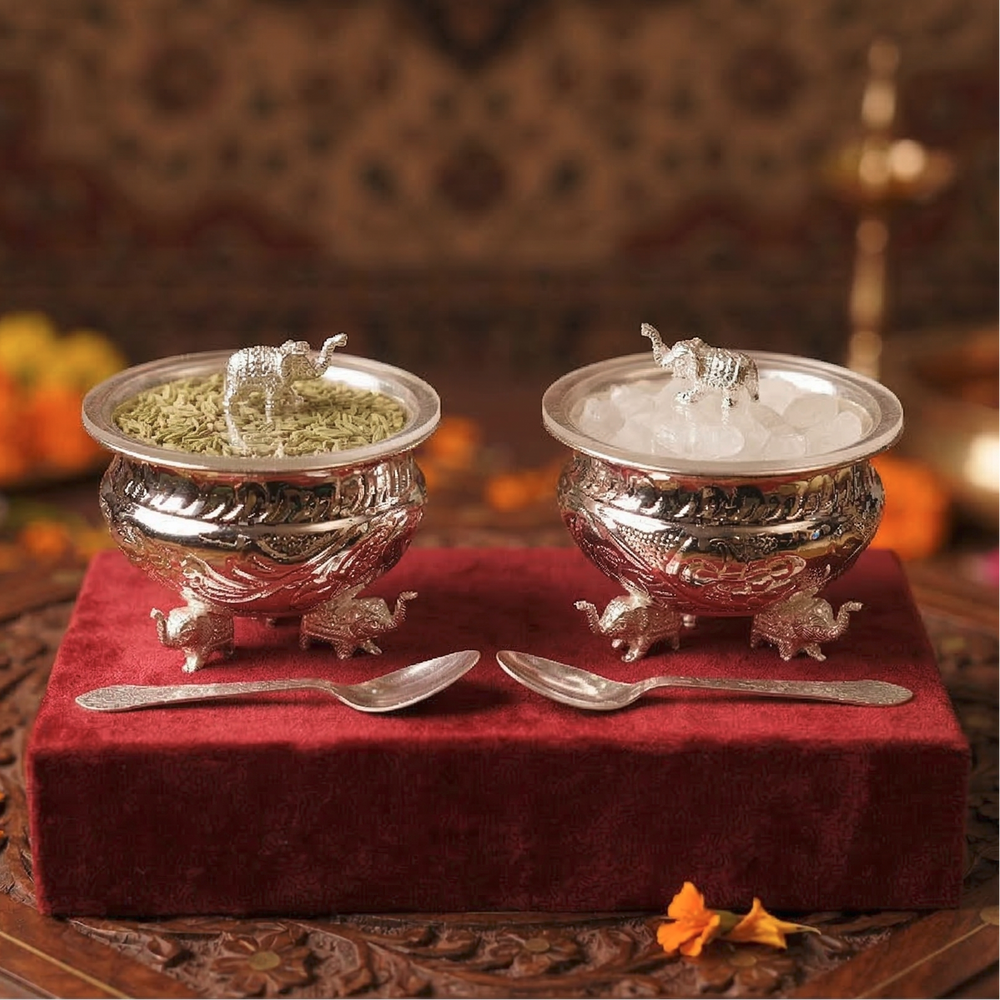 Two silver containers with lids featuring animal sculptures on a red cloth, accompanied by two spoons.