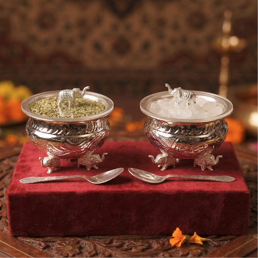Two silver containers with lids featuring animal sculptures on a red cloth, accompanied by two spoons.