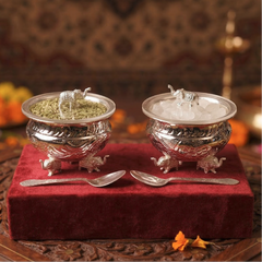 Two silver containers with lids featuring animal sculptures on a red cloth, accompanied by two spoons.