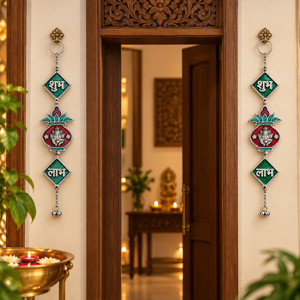 Two decorative hanging wall ornaments with metallic rings, green diamond-shaped panels with white Devanagari text, and colorful floral motifs are displayed on either side of a carved wooden doorway in a warmly lit interior.