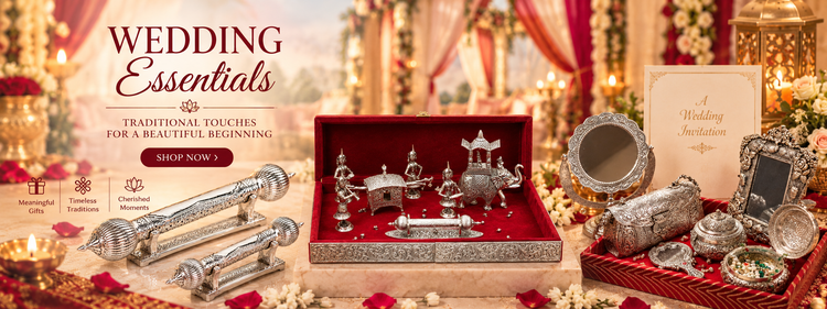 A styled wedding essentials display featuring silver-toned traditional jewelry and accessories, including a silver mirror, silver earrings, rings, a small silver frame, and a silver cylindrical ornament, arranged around an open red velvet box with miniature silver figurines. The scene is decorated with warm lighting, draped fabrics, candles, and floral elements, with text reading 'WEDDING Essentials' on the left.