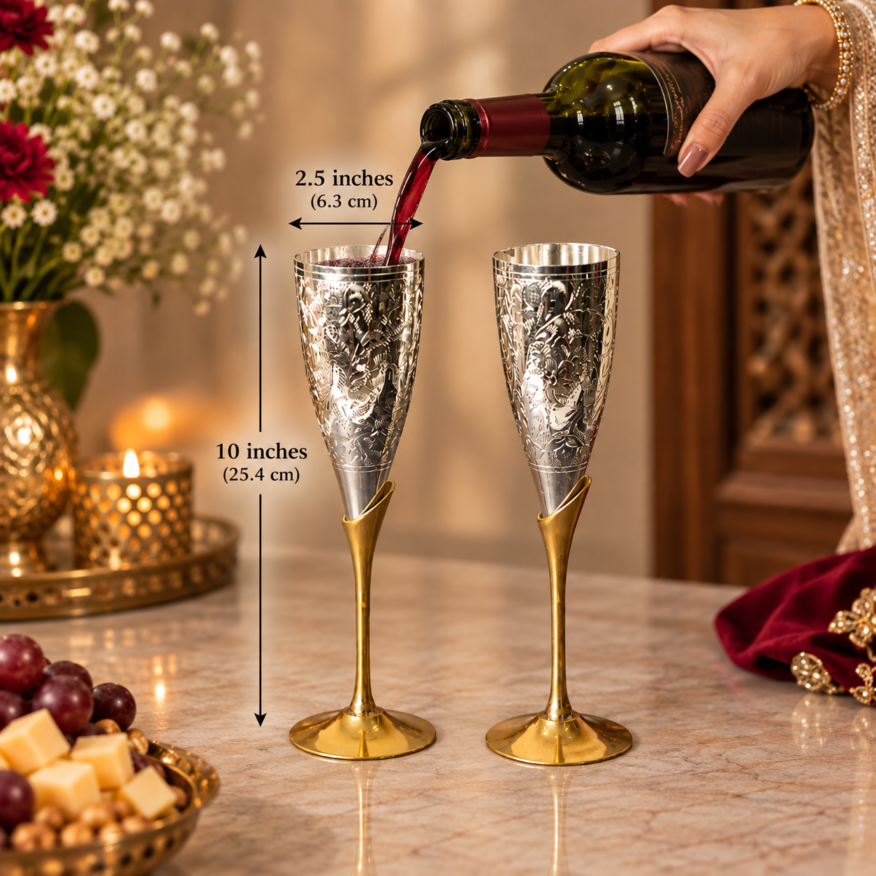 Two ornate champagne flutes with silver engraved bodies and gold stems are shown on a marble table, with a hand pouring red wine from a bottle. Black measurement arrows and text indicate the flutes are 10 inches tall with 2.