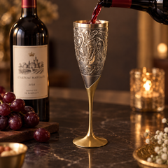 An ornate silver-toned engraved wine flute with a gold-toned stem and base, being filled with red wine from a bottle, set on a dark marble surface with a wine bottle, grapes, and a lit candle in the background.